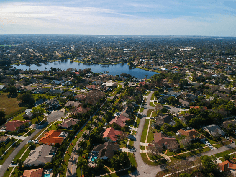Aerial view of a suburban neighborhood with tree-lined streets, single-family homes, and a lake in the background under a clear sky.