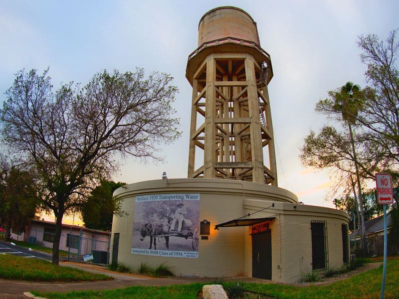 A tall, tan historic water tower stands behind a round brick building with a mural. Leafless trees and a No Parking sign are nearby, under a cloudy sky at sunset.