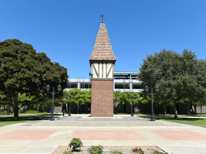 A small brick tower with a peaked roof stands in the center of a courtyard, surrounded by trees, lampposts, and a clear blue sky overhead.