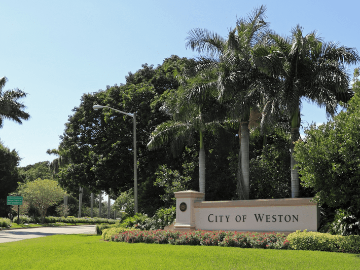 A large white sign reading City of Weston stands among lush green grass, colorful flowers, palm trees, and dense foliage under a clear blue sky.