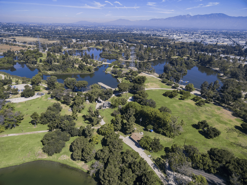 Aerial view of a large park with several lakes, green lawns, and trees, with a cityscape and mountains visible in the background under a clear blue sky.