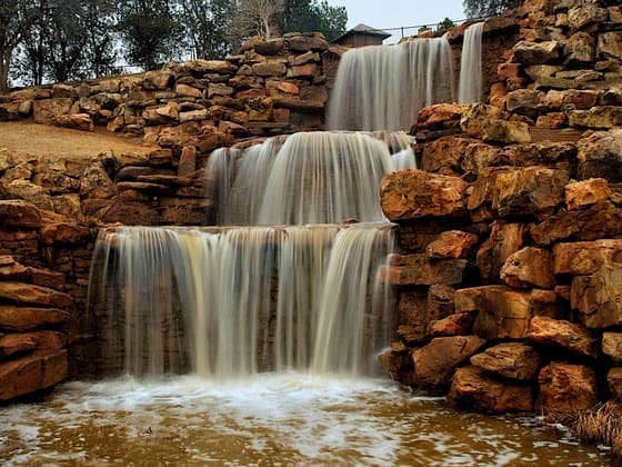 A tiered artificial waterfall cascades over reddish-brown rocks into a small pool, surrounded by stones and sparse trees in the background.