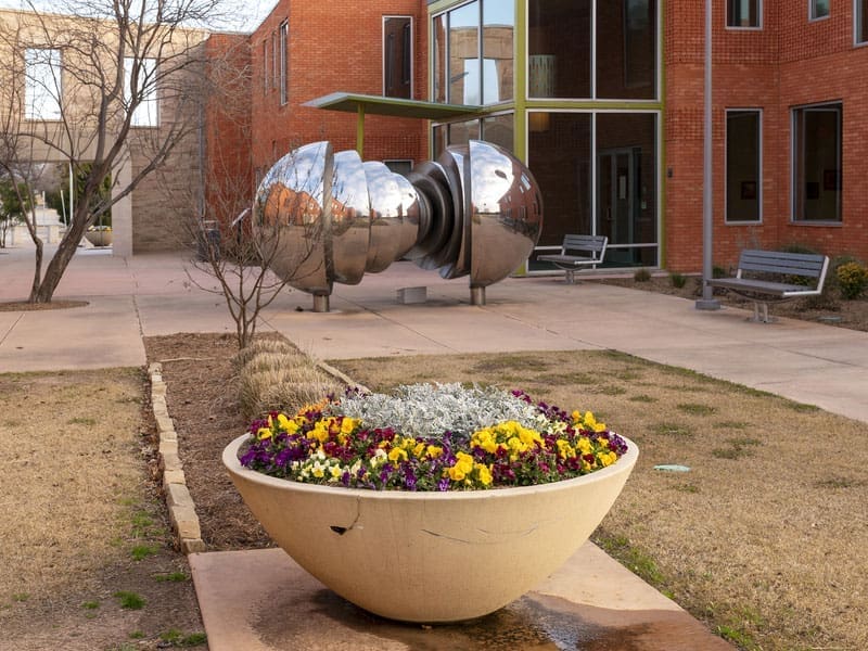 A large bowl-shaped planter with colorful flowers sits in a courtyard, with a modern silver sculpture and brick buildings in the background. Benches and bare trees are also visible.