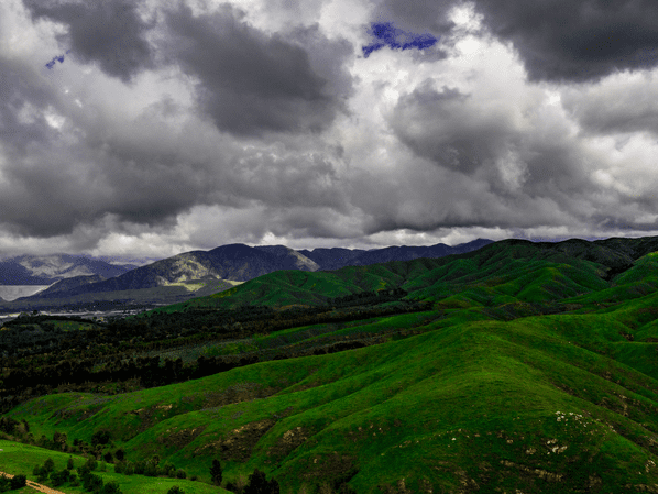 Green rolling hills under dramatic, cloudy skies with mountains in the background and patches of sunlight breaking through the clouds, creating a vibrant, contrasting landscape.