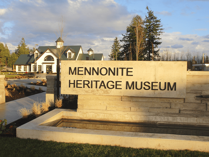 A large sign reading Mennonite Heritage Museum stands in front of a modern building with green roofs, surrounded by trees and landscaping under a partly cloudy sky.