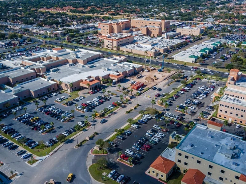 Aerial view of a large medical complex with multiple beige and white buildings, several busy parking lots filled with cars, and surrounding roads in a suburban area. Trees and other buildings are visible in the background.