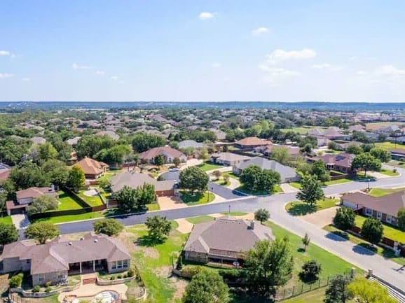 Aerial view of a suburban neighborhood with single-family homes, green lawns, trees, and winding streets under a clear blue sky.