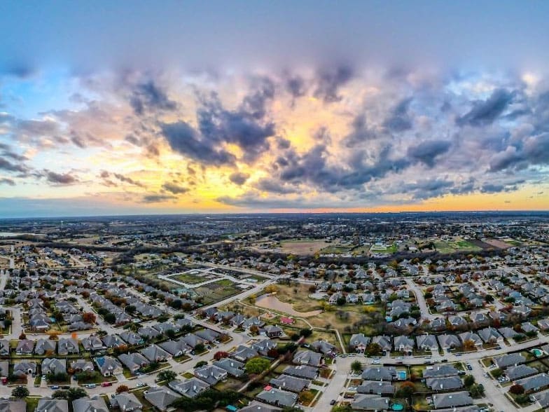 Aerial view of a suburban neighborhood with many houses, roads, and green spaces at sunset. The sky is filled with dramatic clouds illuminated by the colorful setting sun.