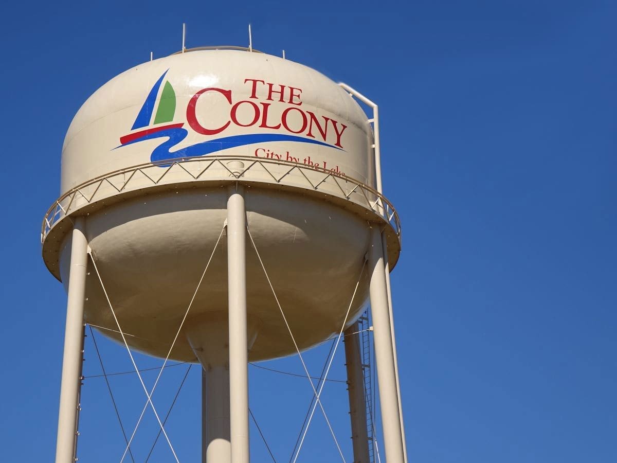 A tall water tower against a clear blue sky displays the words “The Colony” with a sailboat logo and the phrase “City by the lake” in red lettering.
