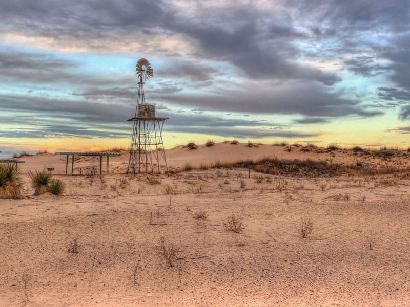 A lone windmill stands in a sandy desert landscape with sparse vegetation, under a cloudy sky at sunset.